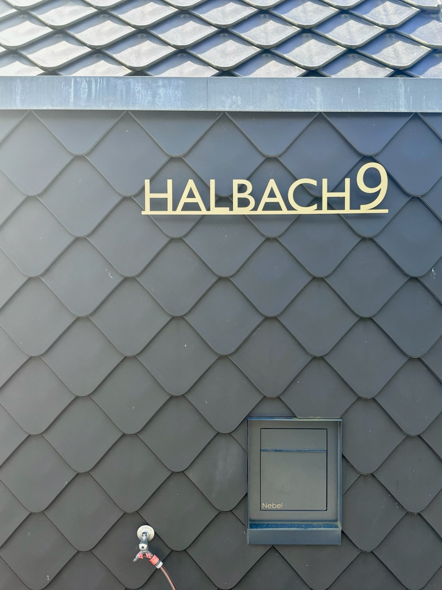 Detail Fassade Schwarze Dachziegel mit goldener Beschriftung "HALBACH 9" über einem kleinen Fenster.