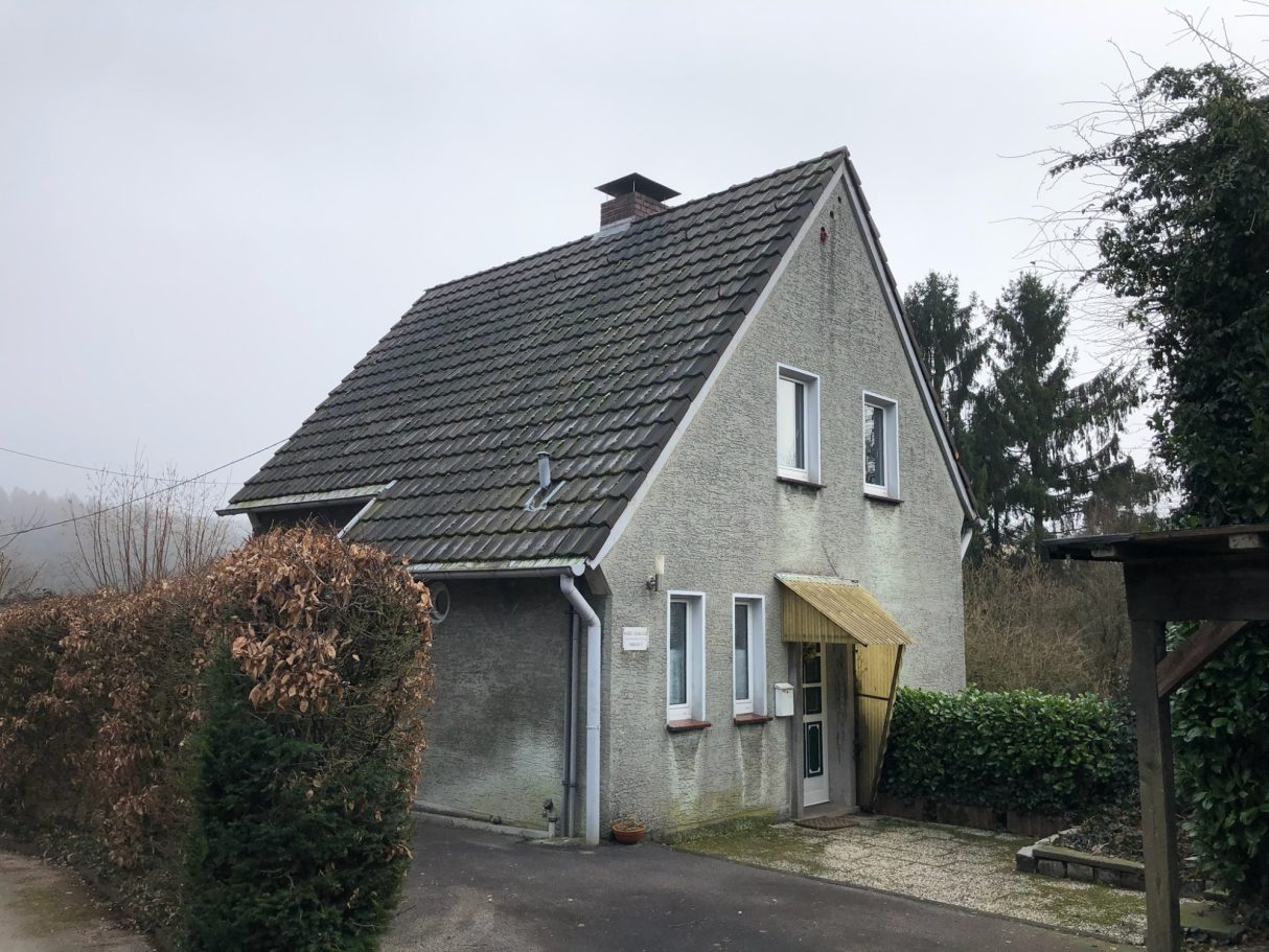 Bestandshaus Zweistöckiges Haus mit grauer Fassade und Satteldach, umgeben von Büschen.