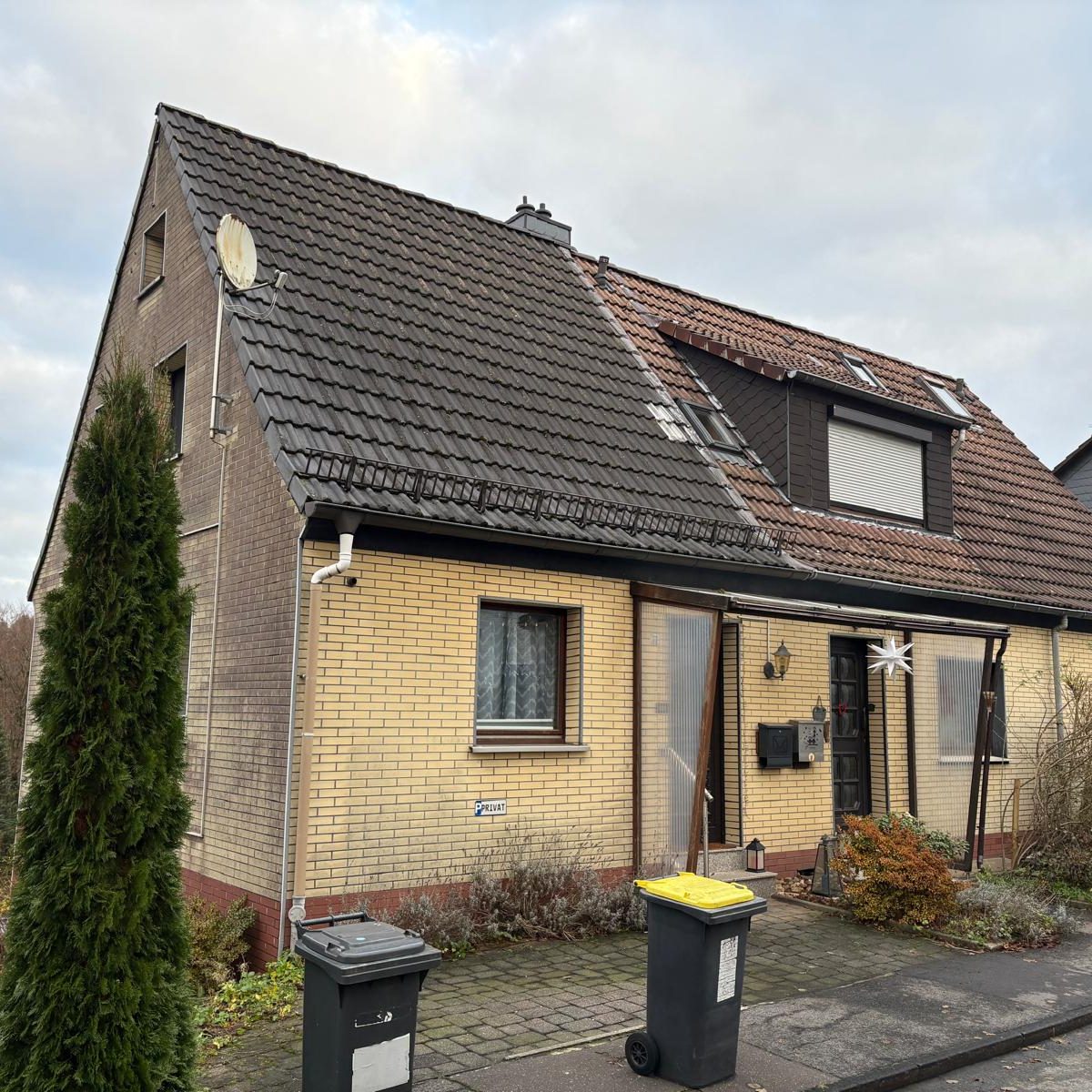 Projekt S31 Doppelhaaushälfte mit grauem Dach, gelber Fassade und zwei Mülltonnen vor der Tür.