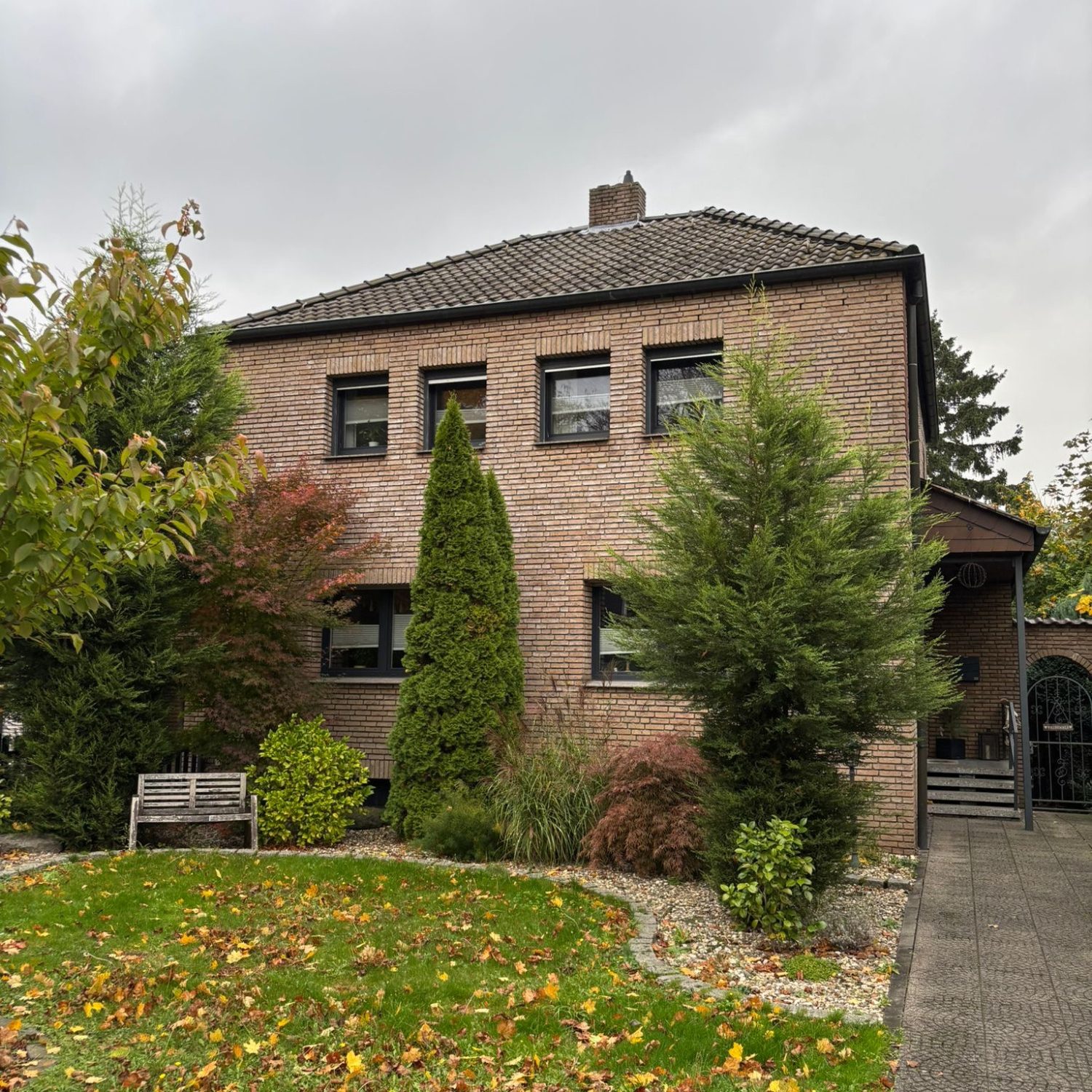 Projekt F7 Zweistöckiges, ziegelrotes Wohnhaus mit Garten und Bänken, umgeben von Bäumen.