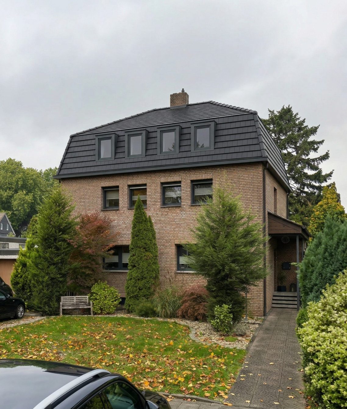 Visualisierung – Dachumbau mit Mansarddach (KI-gestützte Darstellung) Zweistöckiges Haus mit schwarzem Dach und umgebendem Garten, umgeben von Bäumen.