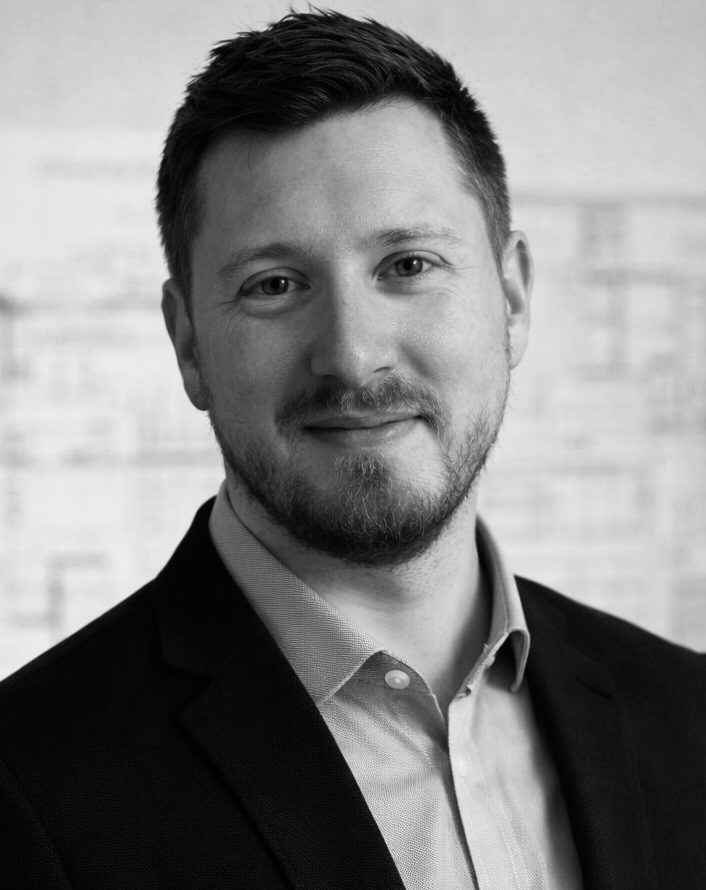 Architekt Michael Nebel Ein lächelnder Mann mit kurzem Bart, in einem dunklen Anzug und leicht geöffnetem Hemd.